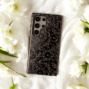 May include: A black phone case with a decorative pattern of moons, stars, and floral elements in a light pink color. The case has a glossy finish and a camera module with multiple lenses. The case is surrounded by white flowers.