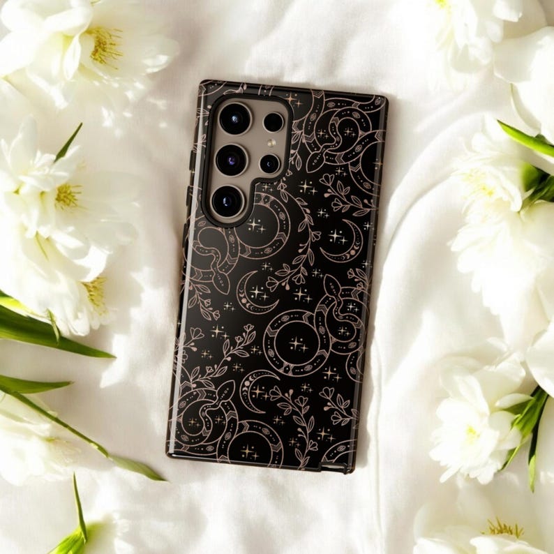 May include: A black phone case with a decorative pattern of moons, stars, and floral elements in a light pink color. The case has a glossy finish and a camera module with multiple lenses. The case is surrounded by white flowers.