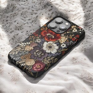 K&ouml;nnte beinhalten: Eine schwarze Smartphone-H&uuml;lle mit einem lebendigen Blumenmuster. Die H&uuml;lle zeigt eine detaillierte Anordnung von roten, wei&szlig;en, lila und gelben Blumen mit gr&uuml;nen Bl&auml;ttern. Die Kameralinsen des Telefons sind oben sichtbar.