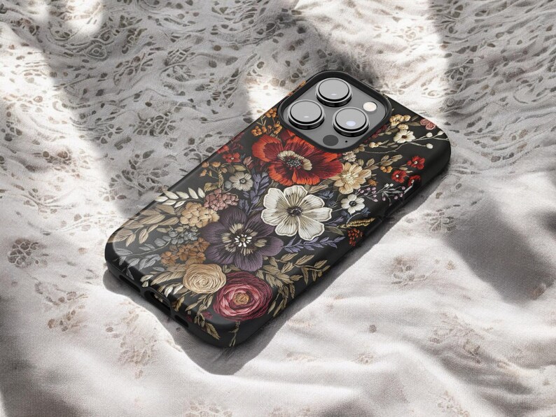 K&ouml;nnte beinhalten: Eine schwarze Smartphone-H&uuml;lle mit einem lebendigen Blumenmuster. Die H&uuml;lle zeigt eine detaillierte Anordnung von roten, wei&szlig;en, lila und gelben Blumen mit gr&uuml;nen Bl&auml;ttern. Die Kameralinsen des Telefons sind oben sichtbar.