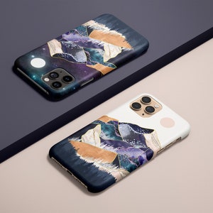 May include: Two smartphone cases with a celestial mountain design. The cases feature a dark blue background with purple, gold, and white accents. One case is on a dark blue surface, the other on a light pink surface.