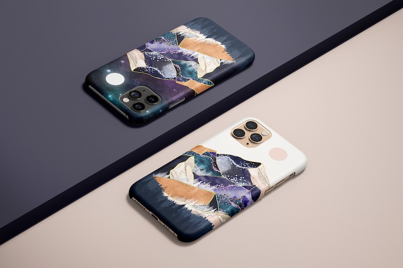 May include: Two smartphone cases with a celestial mountain design. The cases feature a dark blue background with purple, gold, and white accents. One case is on a dark blue surface, the other on a light pink surface.