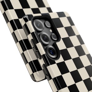 May include: A phone case with a black and cream checkerboard pattern. The case has a glossy finish and features a camera module with multiple lenses and buttons on the side. The design is a classic, retro style.