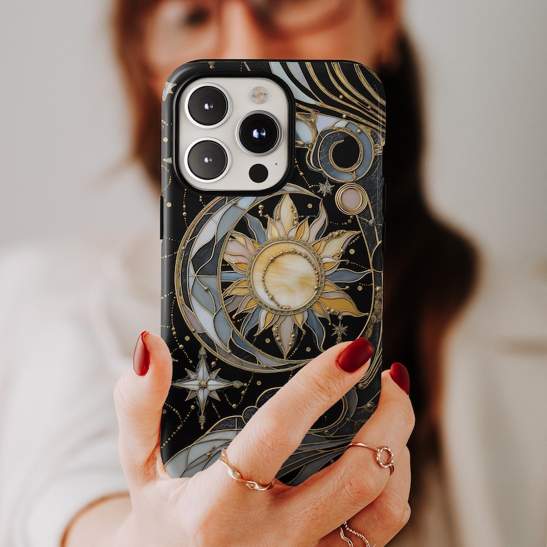 Pu&ograve; includere: Custodia per telefono nera con un design celeste con sole, luna e stelle. La custodia ha un effetto vetrata con accenti dorati e blu. Il design include una stella e una luna crescente. La custodia del telefono &egrave; tenuta in mano.