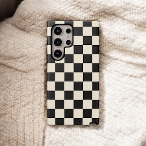 May include: A phone case with a black and cream checkerboard pattern. The case has a camera cutout with multiple lenses and is resting on a textured, off-white surface.
