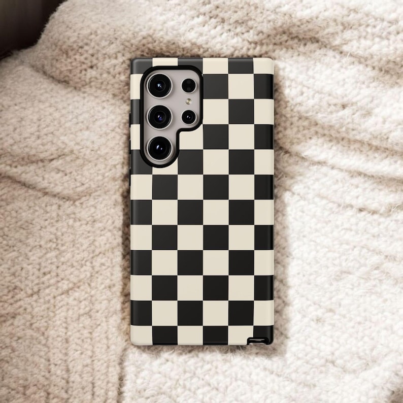 May include: A phone case with a black and cream checkerboard pattern. The case has a camera cutout with multiple lenses and is resting on a textured, off-white surface.