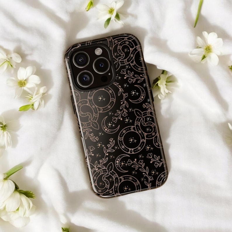 May include: Black phone case with a decorative pattern of snakes, crescent moons, and floral elements in a light brown color. The case has a glossy finish and a camera cutout. White flowers are scattered around the phone case on a white surface.