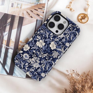 May include: A navy blue phone case with a floral pattern in white and light blue. The case is on a white surface with a gold necklace and a picture of a building in the background.