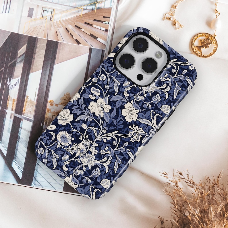 May include: A navy blue phone case with a floral pattern in white and light blue. The case is on a white surface with a gold necklace and a picture of a building in the background.
