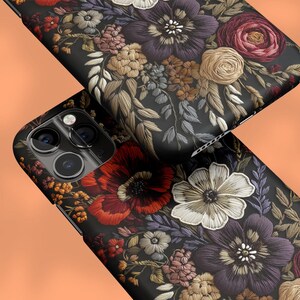 K&ouml;nnte beinhalten: Zwei schwarze Handyh&uuml;llen mit matter Oberfl&auml;che, die ein detailliertes Blumenmuster aufweisen. Das Design umfasst Blumen im Stickstil in Rot-, Lila-, Wei&szlig;- und Beiget&ouml;nen. Die H&uuml;llen sind vor einem orangefarbenen Hintergrund abgewinkelt.