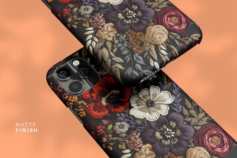 K&ouml;nnte beinhalten: Zwei schwarze Handyh&uuml;llen mit matter Oberfl&auml;che, die ein detailliertes Blumenmuster aufweisen. Das Design umfasst Blumen im Stickstil in Rot-, Lila-, Wei&szlig;- und Beiget&ouml;nen. Die H&uuml;llen sind vor einem orangefarbenen Hintergrund abgewinkelt.