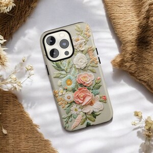 iPhone case with an intricate floral embroidery. lying on top of a soft fabric background with delicate flowers and leaves. The case features focus stacking, cottagecore aesthetics, vintage style