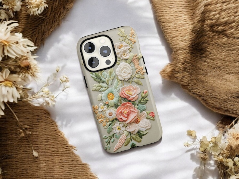 iPhone case with an intricate floral embroidery. lying on top of a soft fabric background with delicate flowers and leaves. The case features focus stacking, cottagecore aesthetics, vintage style
