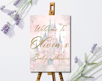 Puede incluir: Un letrero de baby shower sobre un caballete de madera. El letrero presenta una ilustración de acuarela de un castillo rosa con torres azules y el texto "Welcome To Olivia's Baby Shower" en escritura dorada. Ramas de lavanda enmarcan el letrero.