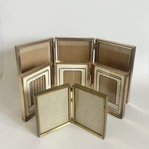 May include: A collection of seven hinged brass picture frames arranged in a tiered display. The frames feature metallic gold-toned edges with a beaded texture, clear glass panels, and off-white matting, creating a multi-panel folding structure.