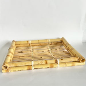 May include: A rectangular bamboo tray with a natural finish. The tray is constructed from multiple bamboo stalks, with a slatted interior and rounded edges. White string secures the frame. The tray is suitable for serving food or as a decorative item.