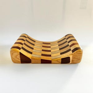 May include: A handcrafted wooden cutting board with a unique, curved design. The board features a checkerboard pattern of light and dark wood tones, including oak and walnut. The board is approximately 12 inches long and 8 inches wide.