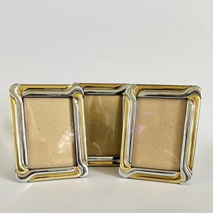 May include: Three rectangular photo frames with a silver and gold-tone metal design. The frames have a curved, layered border and a neutral-colored backing. The frames are arranged side by side on a white surface.