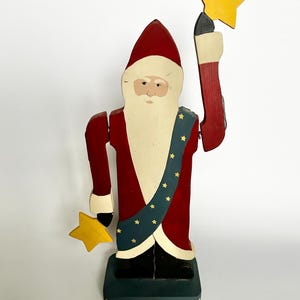 May include: Wooden Santa Claus figurine with a red coat, hat, and arms, holding two yellow stars. The figurine has a cream-colored face and beard, a blue sash with yellow stars, and stands on a green base.