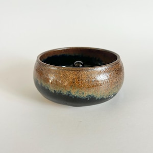 May include: A round ceramic bowl with a glossy finish, featuring a gradient of dark black at the base transitioning into a warm, speckled brown hue toward the rim. A small, smooth, dark spherical object is centered at the bottom of the bowl.