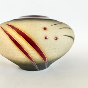 May include: A ceramic vase with a unique design. The vase has a beige body with diagonal red stripes and small red dots. The top and base are dark green, and the interior is a dark brown color. The vase has a rounded, organic shape.