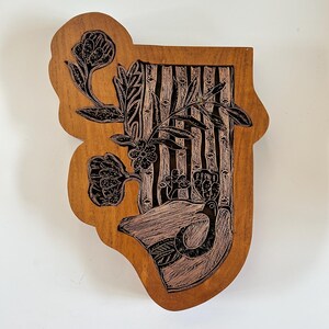 May include: A natural-toned wooden plaque with an irregular, organic shape featuring a black ink-stamped design. The artwork depicts a bird perched near a stylized tree trunk, surrounded by floral elements and vertical lines in a detailed print.