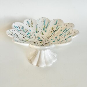 May include: A white ceramic pedestal bowl with a scalloped edge and a flared base. The bowl is decorated with speckled blue, green, and brown accents. The bowl is designed for serving or display.