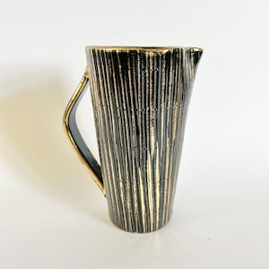 May include: A black and gold pitcher with a textured, vertical striped design. The pitcher has a curved handle and a spout. The gold accents add a touch of elegance to the ceramic pitcher.