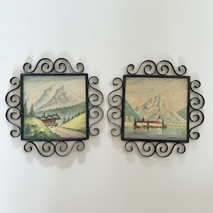 May include: Two square wall plaques featuring landscape paintings of mountain scenes. Each painting is encased in a black metal frame with decorative scrollwork edges. The left plaque shows a house near mountains, while the right shows a building on water.