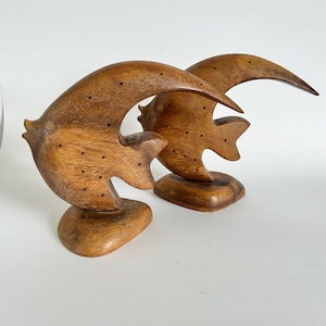 May include: Two carved wooden fish sculptures with a natural brown finish. Each fish features a curved, crescent-like body shape, a smooth surface with visible wood grain, and a series of small, evenly spaced dark dots across the side.