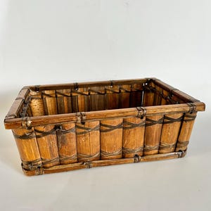 May include: Rectangular bamboo basket with a natural wood finish. The basket is constructed from vertical bamboo poles held together with dark brown woven bands. The rim is reinforced with a thicker bamboo frame.
