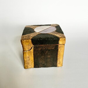 May include: A small, square box with a hinged lid. The box is black with gold-colored edges and a geometric design on the top. The top design features a silver and lavender oval shape. The box appears to be made of a textured material.