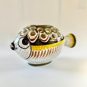 May include: A ceramic fish-shaped vase with a white, cracked glaze. The vase features a dark brown eye, a yellow stripe, and a green top with multiple holes. The tail is yellow and white. The vase is a decorative item.