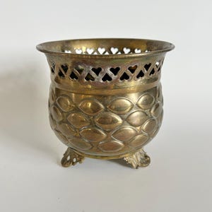 May include: A small, round brass vessel featuring a textured, scale-like pattern on its exterior body. The upper rim is adorned with a decorative cutout design of small hearts, and the base is supported by three ornate, curved metal feet.