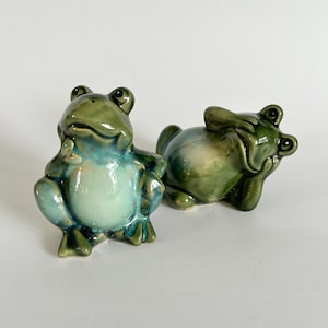 May include: Two ceramic frog figurines. One frog is standing with its hands on its hips, and the other is lying down. The frogs are glazed in shades of green and blue, with a glossy finish. The figurines are decorative.