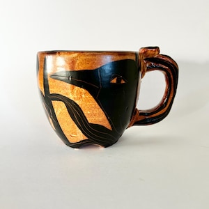 May include: A handmade ceramic mug with a unique design. The mug features a black and copper color scheme with a stylized bird or face design. The handle is also black and copper. The mug is suitable for coffee, tea, or other beverages.