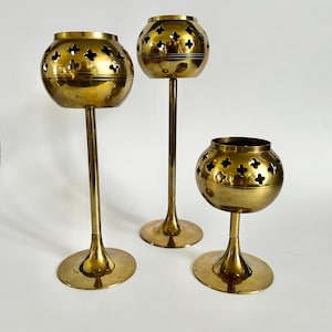 May include: Three brass candle holders of varying heights. Each holder has a round base, a slender stem, and a spherical top with decorative cut-out patterns. The brass has a warm, golden hue, and the design is vintage.