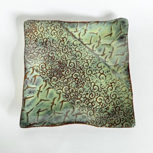 May include: Square ceramic plate with a unique design. The plate features a diagonal split with two distinct patterns. One side has brown swirls, the other has brown bird-like shapes on a green background. The edges are a darker brown.