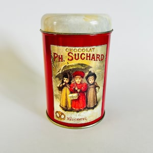 May include: Red metal tin with a white lid. The tin has a label with the text "CHOCOLAT Ph. SUCHARD" and an illustration of three children walking under an umbrella. The label also includes the text "Neuchatel Suisse".