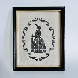 May include: Black and white embroidered picture of a woman in a dress framed in a black frame with a gold trim.