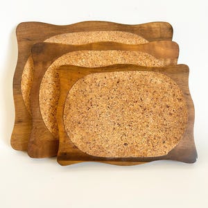 May include: A stack of four wooden trivets with a wavy edge design. Each trivet has a cork center. The wood has a warm, brown tone, and the cork has a speckled, natural appearance. The trivets are arranged in a layered fashion.