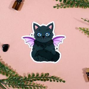 Peut inclure: Autocollant représentant un chat noir aux yeux bleus et aux ailes de chauve-souris violettes. Le chat a une poitrine et une queue touffues et est assis droit. L'autocollant est sur un fond rose avec de petites bouteilles et de la verdure.