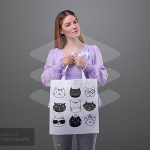 May include: A white tote bag featuring a repeating pattern of illustrated cat faces in various styles. The cats are depicted in black and white with accents of yellow. The bag has white handles and is held by a person.