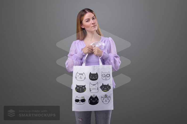 May include: A white tote bag featuring a repeating pattern of illustrated cat faces in various styles. The cats are depicted in black and white with accents of yellow. The bag has white handles and is held by a person.
