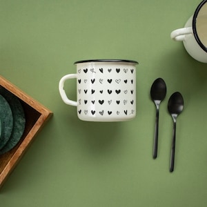 May include: A white enamel mug with a black rim and handle, featuring a pattern of black hearts. The mug is on a green surface with two black spoons and a wooden box with green coasters.