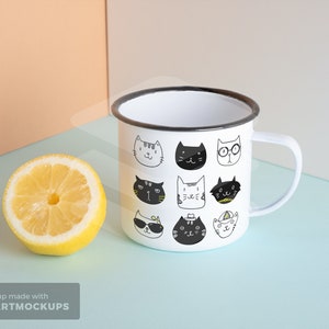 May include: White enamel mug with a black rim and handle, decorated with various cartoon cat faces in black and white. A halved lemon sits beside the mug on a light blue surface.