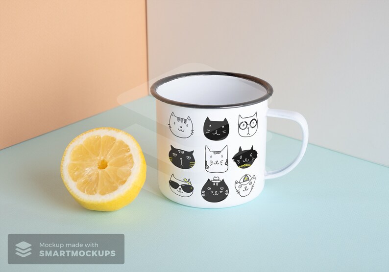 May include: White enamel mug with a black rim and handle, decorated with various cartoon cat faces in black and white. A halved lemon sits beside the mug on a light blue surface.