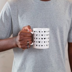 May include: A white ceramic mug with a black and white heart pattern. The mug is being held by a person.