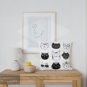 May include: A white square throw pillow with a pattern of illustrated cat faces in black and yellow. The pillow is on a wooden cabinet, with a framed line drawing and a small lamp. A woven basket with lemons is also on the cabinet.