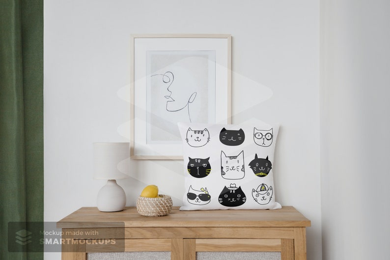 May include: A white square throw pillow with a pattern of illustrated cat faces in black and yellow. The pillow is on a wooden cabinet, with a framed line drawing and a small lamp. A woven basket with lemons is also on the cabinet.